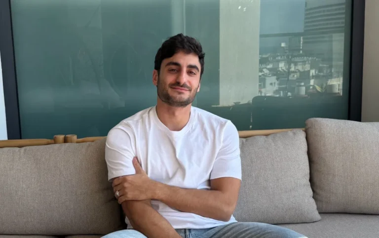 Man in a white t-shirt sitting on a beige couch, smiling slightly, with a cityscape visible through the window behind him.