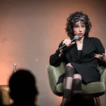 Woman speaking into a microphone while seated on a stage, gesturing with her hand, with an audience silhouette in the fore...