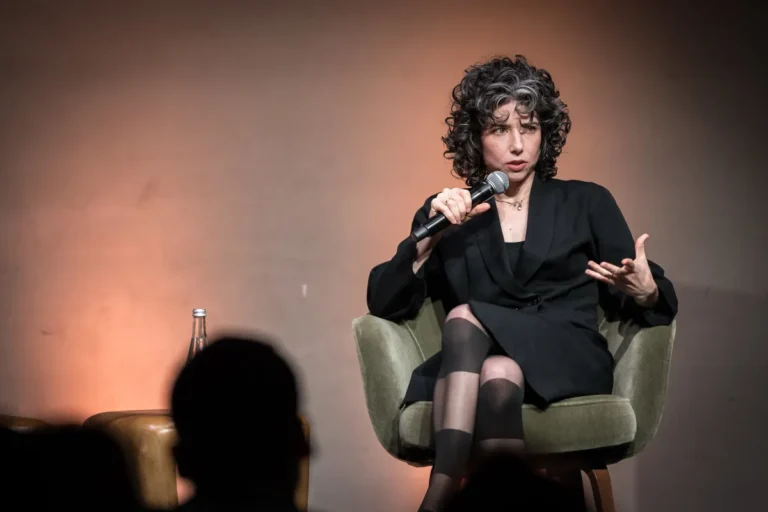 Woman speaking into a microphone while seated on a stage, gesturing with her hand, with an audience silhouette in the fore...