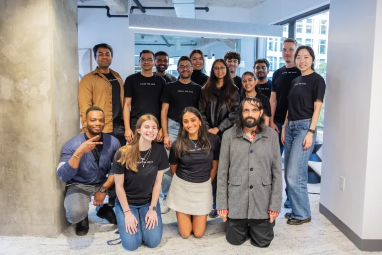 Group of 15 people in casual attire posing indoors, many wearing "Check The Data" shirts, smiling in an office setting.