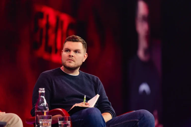 Man seated on stage holding a notebook and pen, with a red backdrop and blurred figure on screen in the background.