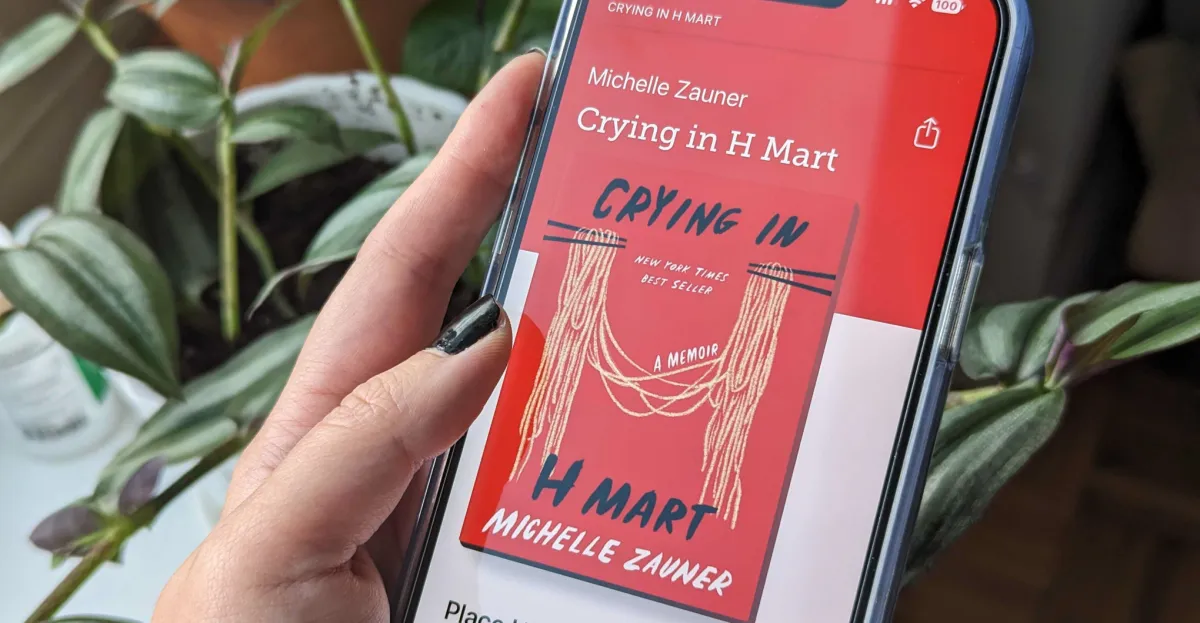 Hand holding a smartphone displaying "Crying in H Mart" book cover with plants in the background.