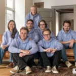 Seven people wearing matching light blue hoodies sit and kneel on a sofa and floor, with a camera on the carpet in front.