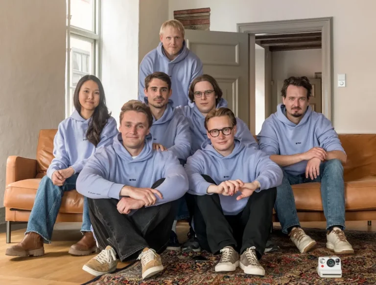 Seven people wearing matching light blue hoodies sit and kneel on a sofa and floor, with a camera on the carpet in front.
