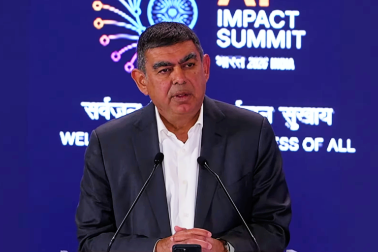 Man in a suit speaking at a podium with "AI Impact Summit" displayed on a screen behind him.