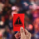 Hand holding a red card with a warning symbol, blurred colorful crowd in the background.