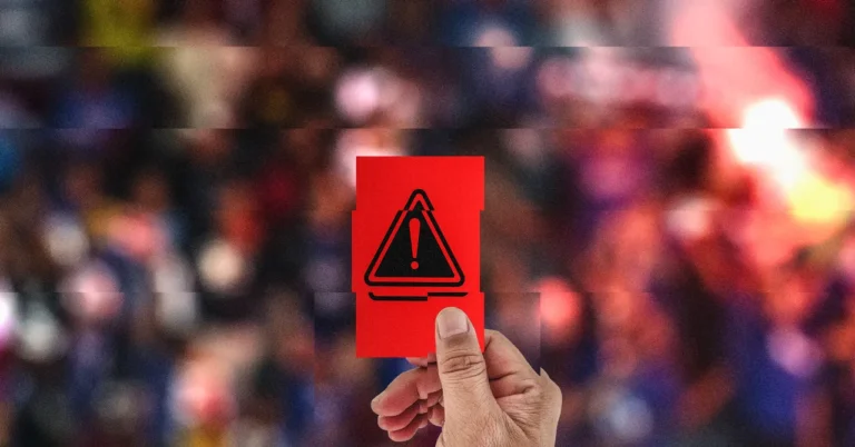 Hand holding a red card with a warning symbol, blurred colorful crowd in the background.