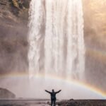 Person standing with arms raised in front of a large waterfall, with two rainbows visible in the mist.