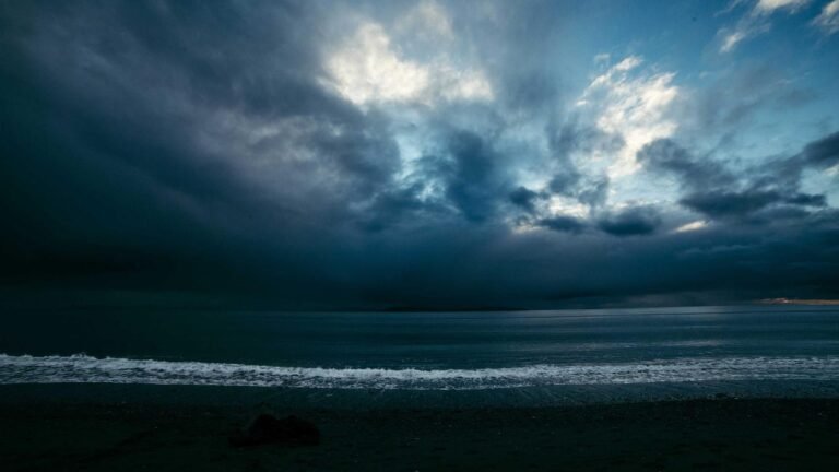 Dark storm clouds over a calm ocean with waves gently hitting a sandy shore, under a partially illuminated sky.