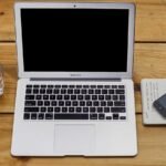 Open laptop on wooden table next to a glass of water, a notebook with sketches, and a smartphone resting on the notebook.