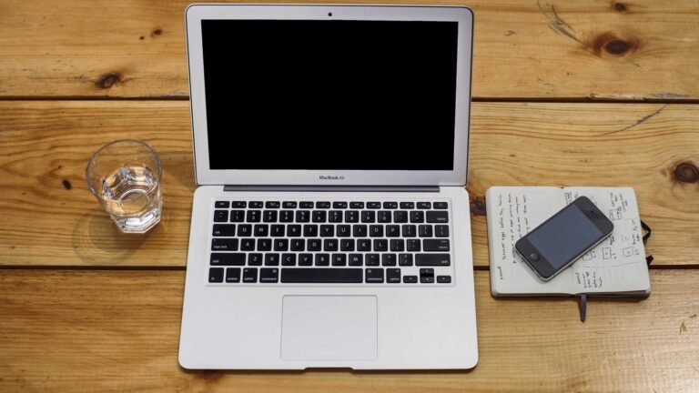 Open laptop on wooden table next to a glass of water, a notebook with sketches, and a smartphone resting on the notebook.