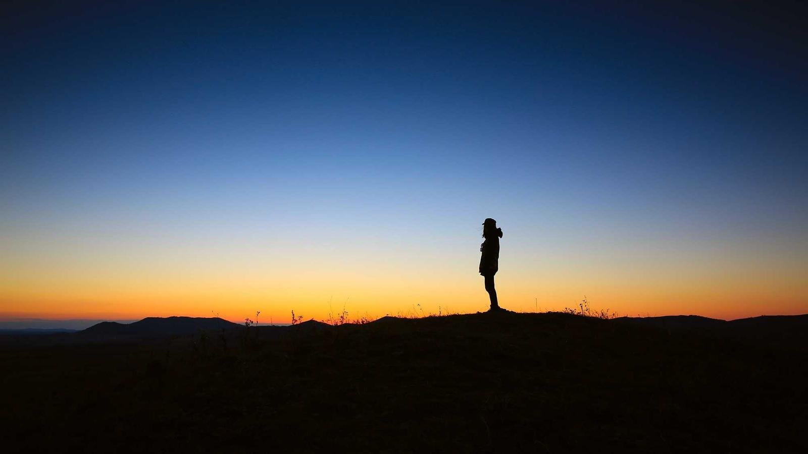Silhouette of a person standing on a hill at sunset with a clear sky transitioning from orange to deep blue.
