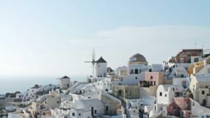 White buildings and windmills on a hillside in Santorini, Greece, with a Greek flag visible, set against the sea and sky.