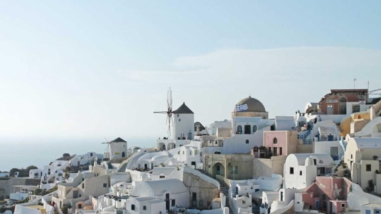 White buildings and windmills on a hillside in Santorini, Greece, with a Greek flag visible, set against the sea and sky.