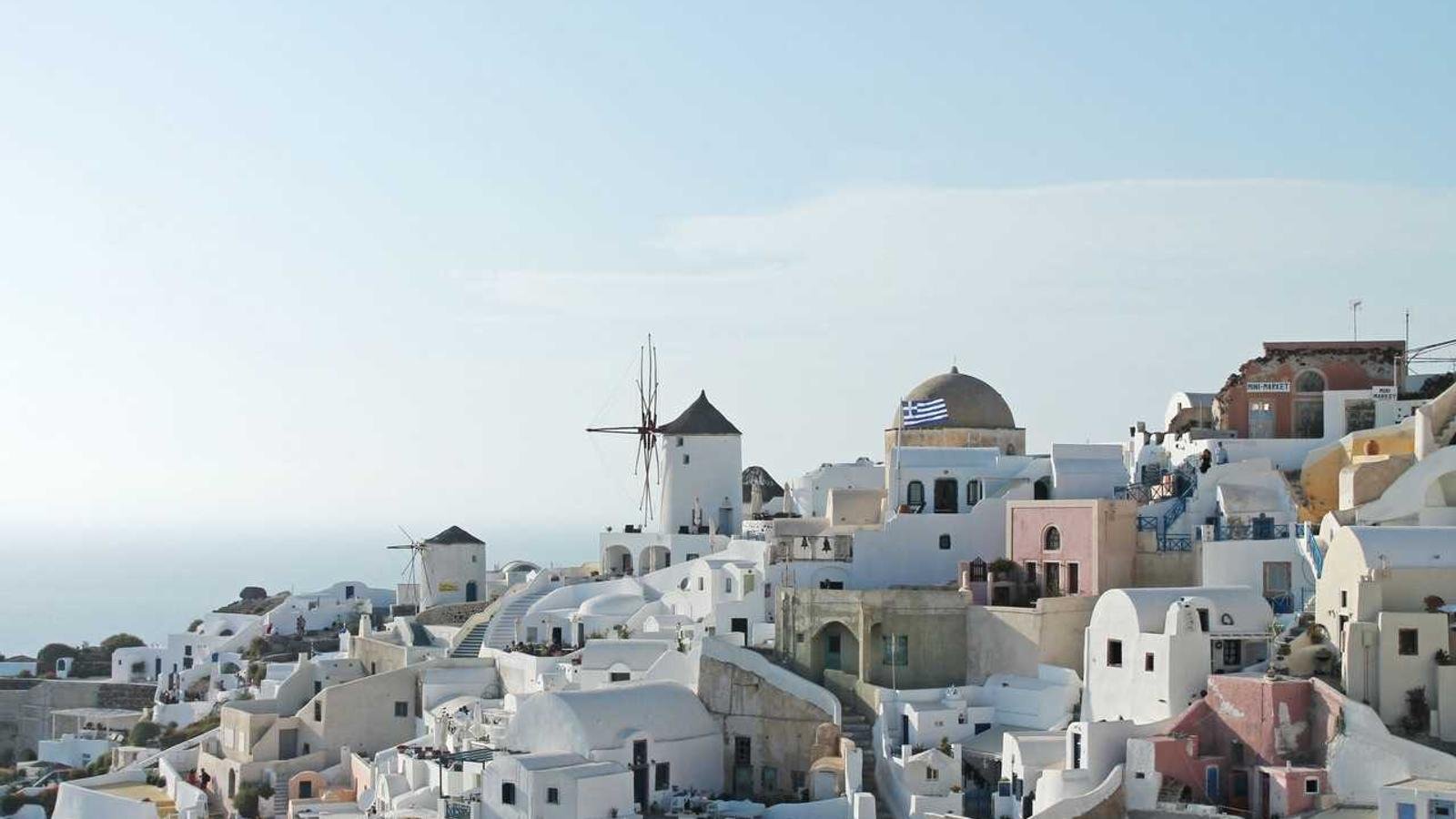 White buildings and windmills on a hillside in Santorini, Greece, with a Greek flag visible, set against the sea and sky.