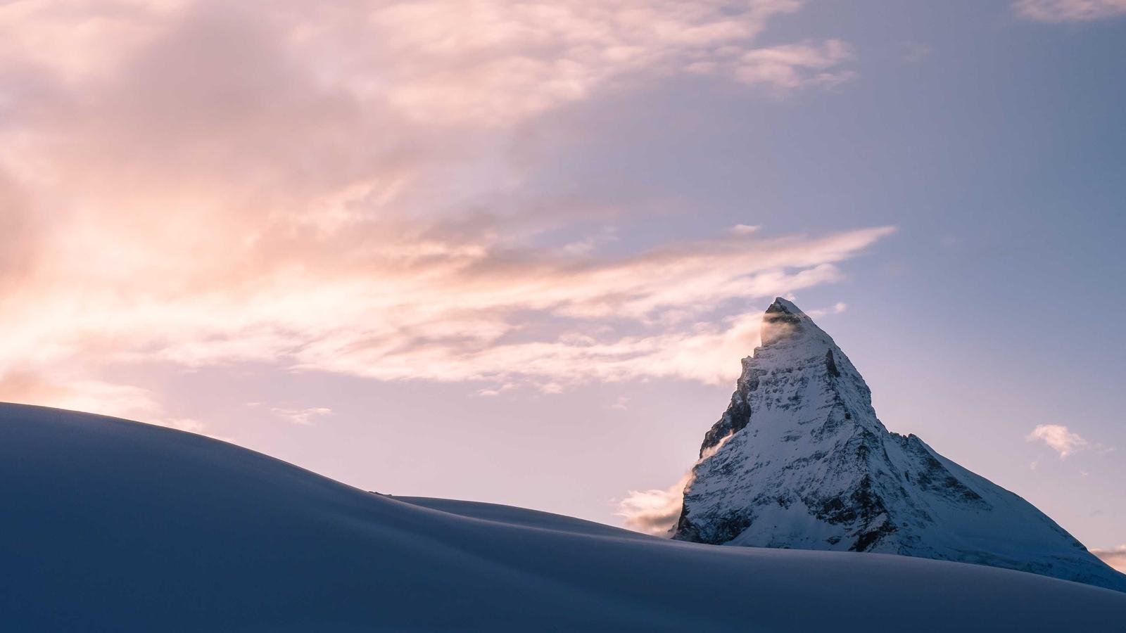 Snow-covered mountain peak under a pink-hued sky with soft clouds, partially illuminated by sunset or sunrise light.