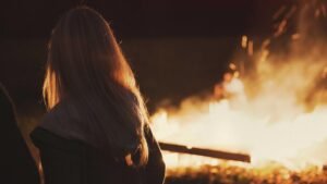 Person with long hair facing a large outdoor fire at night, with sparks flying and a blurred figure in the background.