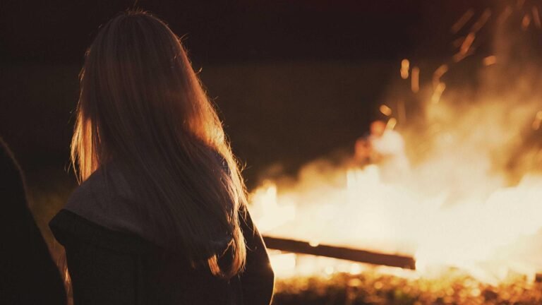 Person with long hair facing a large outdoor fire at night, with sparks flying and a blurred figure in the background.