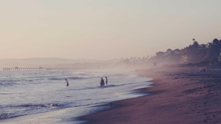 People standing in shallow ocean water at a misty beach, with a pier and cliffs in the background during sunset.