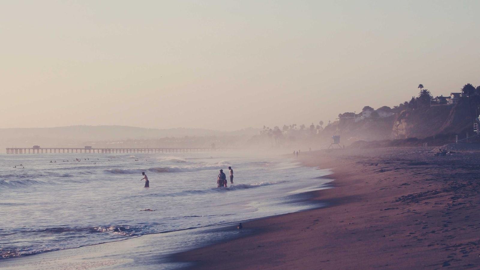People standing in shallow ocean water at a misty beach, with a pier and cliffs in the background during sunset.