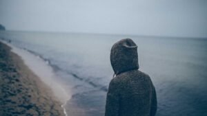 Person in a hooded sweater stands on a deserted beach, facing the calm sea under a gray, overcast sky.