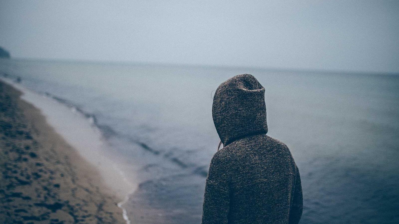 Person in a hooded sweater stands on a deserted beach, facing the calm sea under a gray, overcast sky.