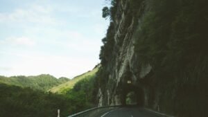 Road passing through a tunnel carved into a rocky hillside, surrounded by lush greenery under a clear sky.