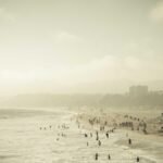 People enjoying a crowded beach with gentle waves under a hazy sky; buildings and mountains visible in the background