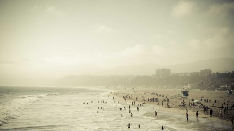 People enjoying a crowded beach with gentle waves under a hazy sky; buildings and mountains visible in the background