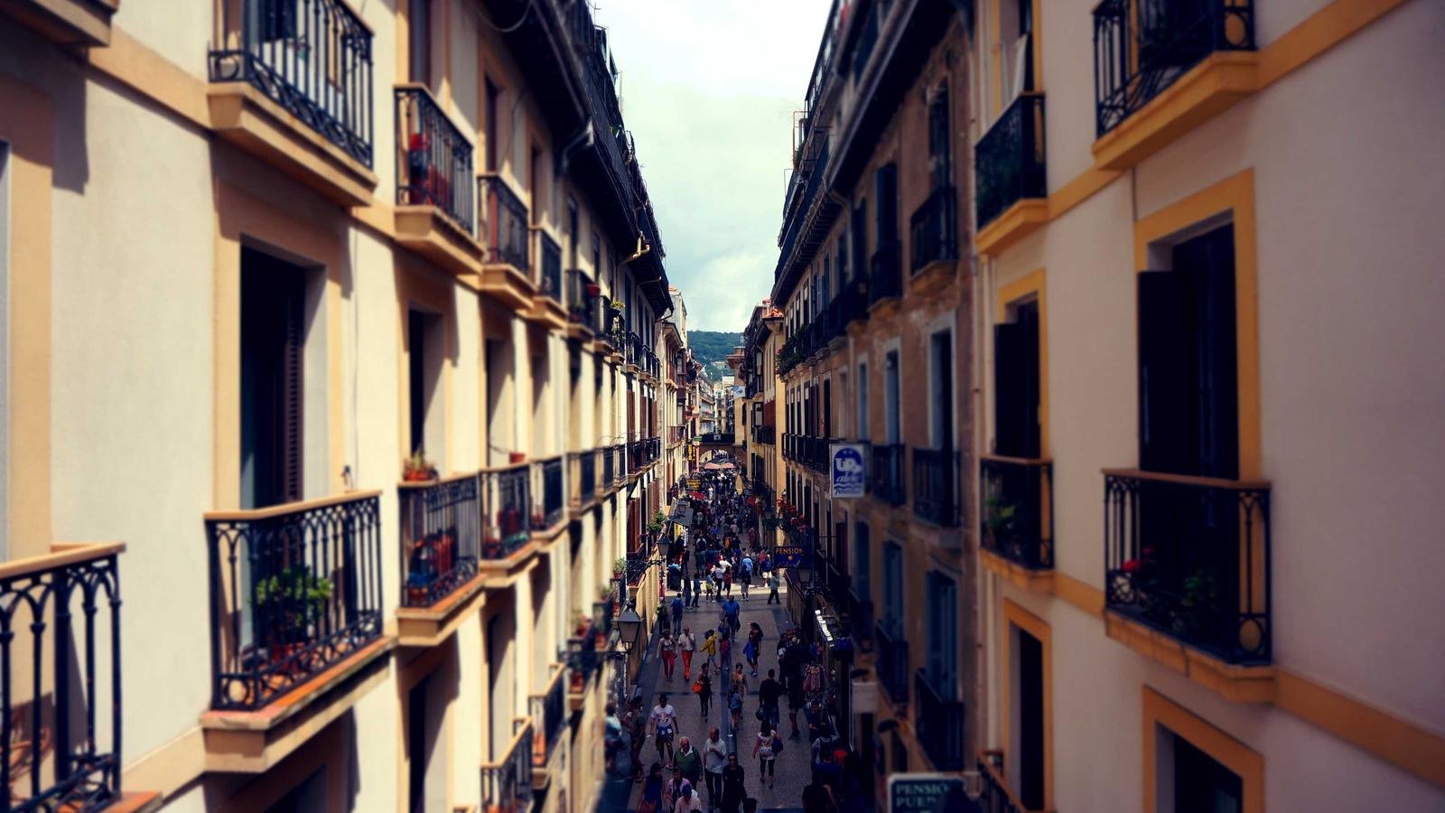 Narrow street lined with buildings featuring balconies, bustling with people walking, some signs visible along the way