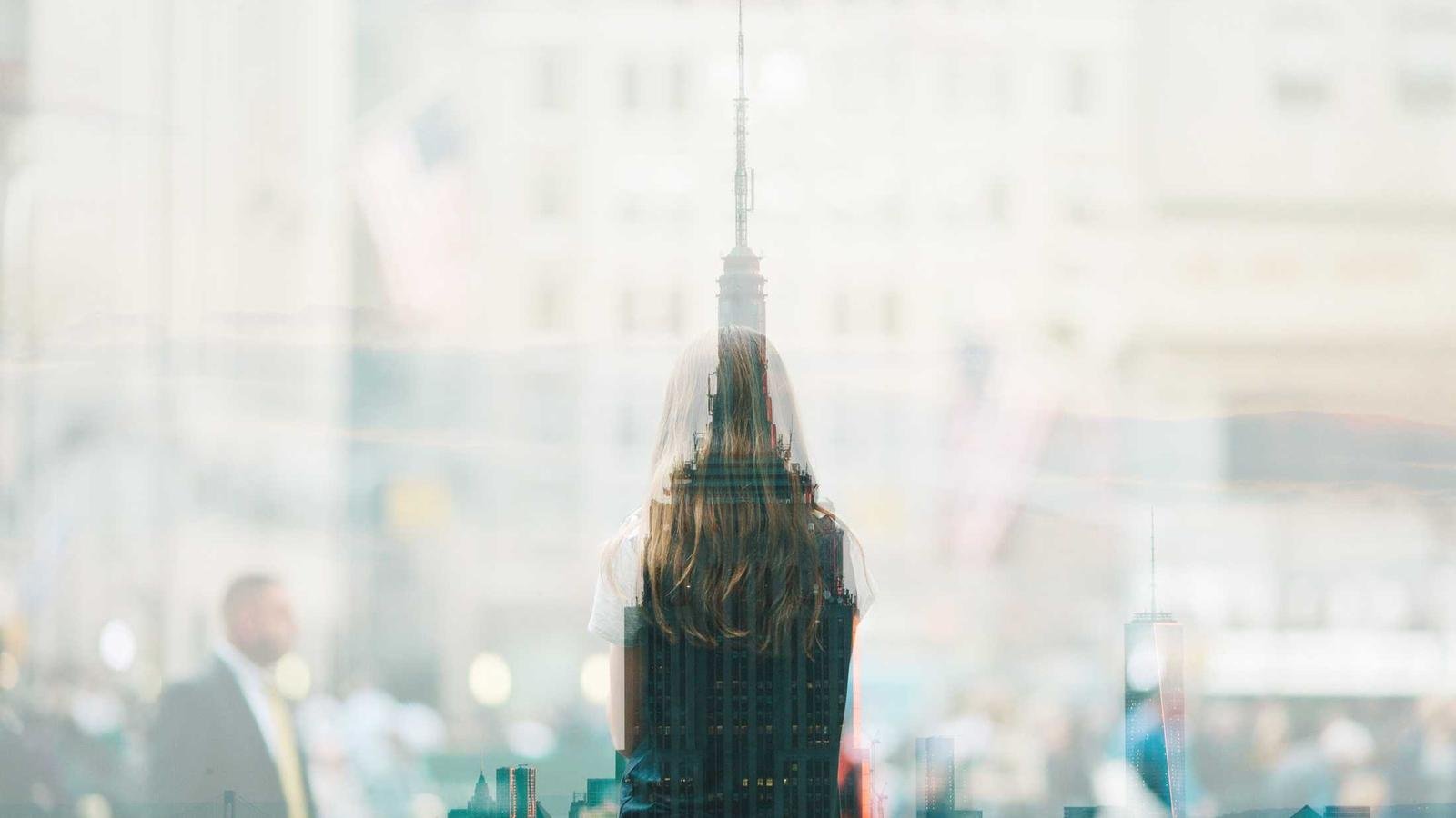 Double exposure of a woman facing away and a cityscape with skyscrapers, blurred crowd in the background.