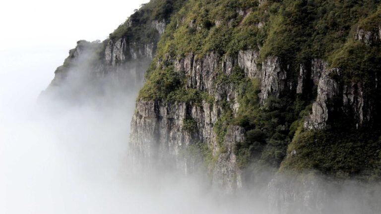 Steep, green-covered rocky cliffs partially obscured by mist, against a white sky backdrop.