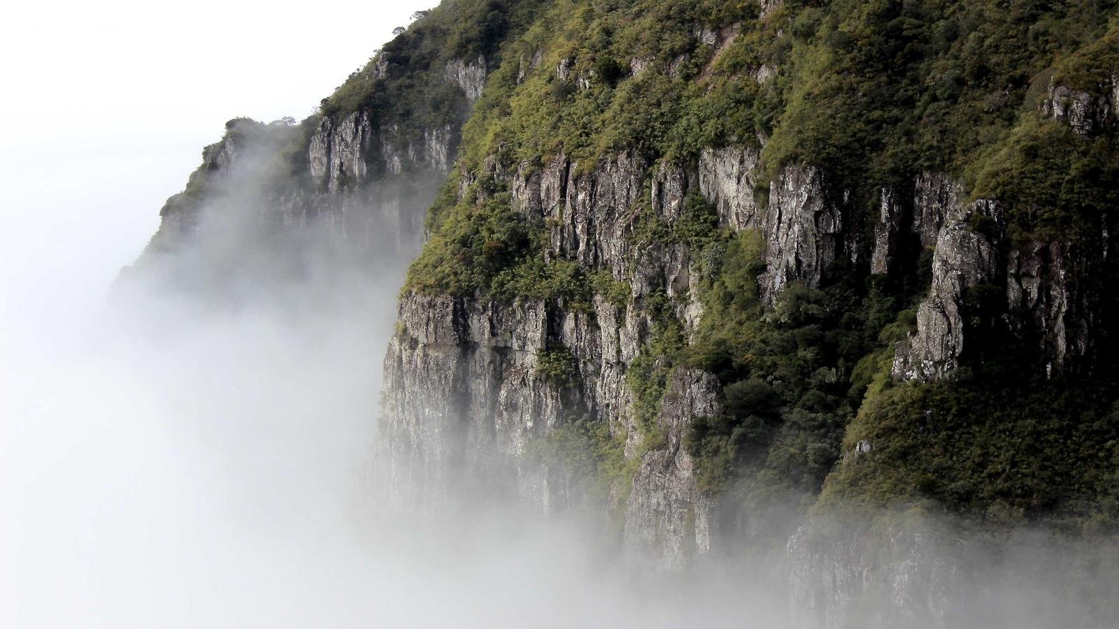 Steep, green-covered rocky cliffs partially obscured by mist, against a white sky backdrop.