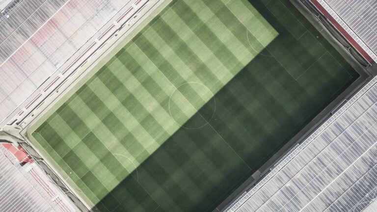 Aerial view of an empty soccer stadium with a green field and striped grass, surrounded by empty seats and a large shadow.