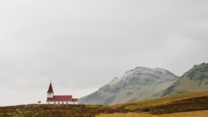 A small white church with a red roof on a grassy hill, mountains in the background under a cloudy sky.