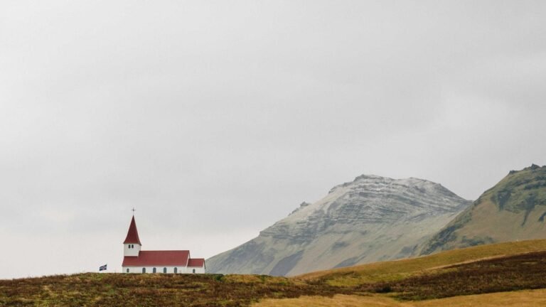 A small white church with a red roof on a grassy hill, mountains in the background under a cloudy sky.