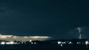 Dark cityscape with distant lightning strike illuminating the night sky, scattered lights dot the skyline.