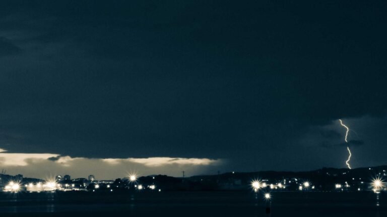 Dark cityscape with distant lightning strike illuminating the night sky, scattered lights dot the skyline.