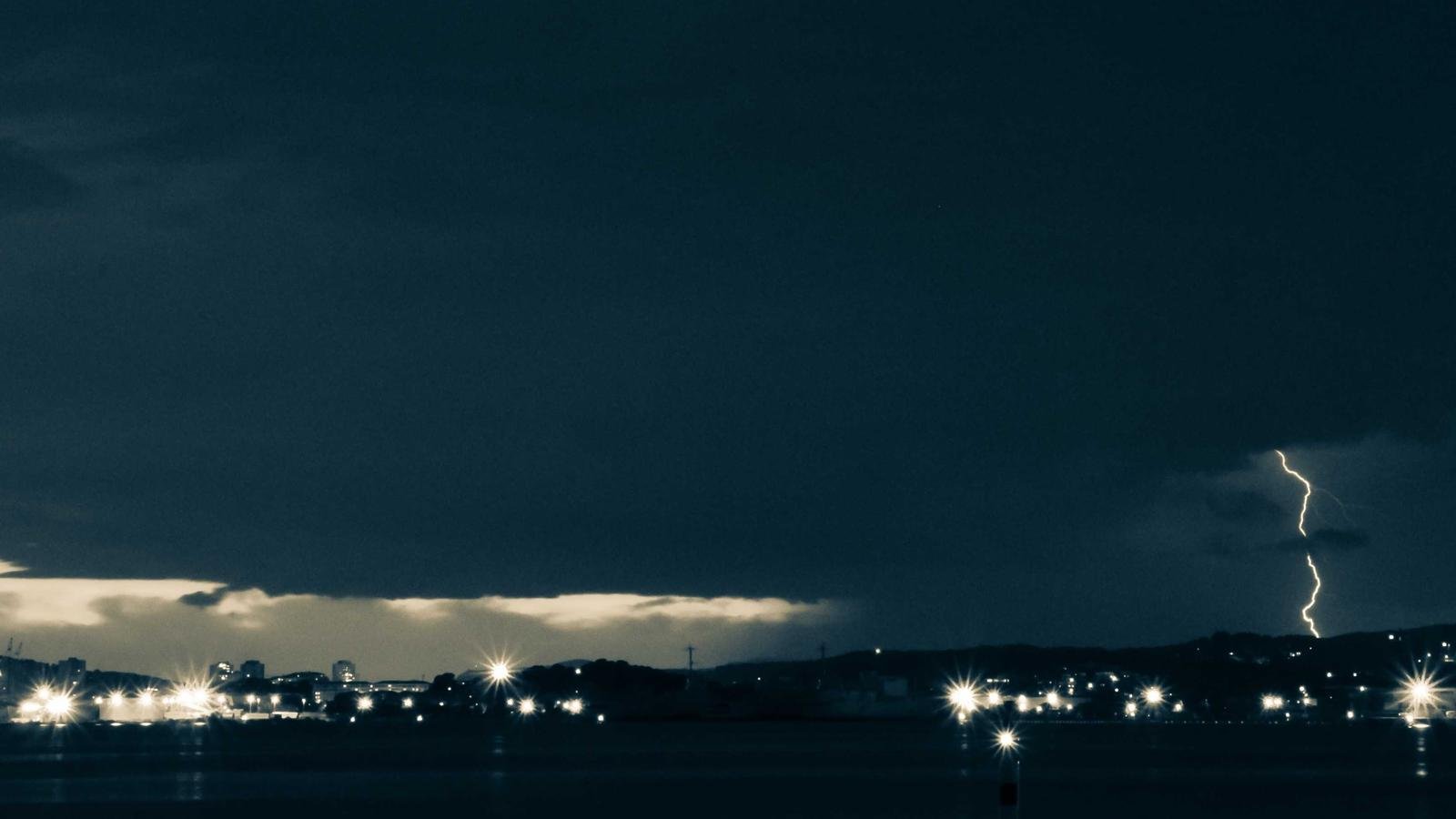 Dark cityscape with distant lightning strike illuminating the night sky, scattered lights dot the skyline.