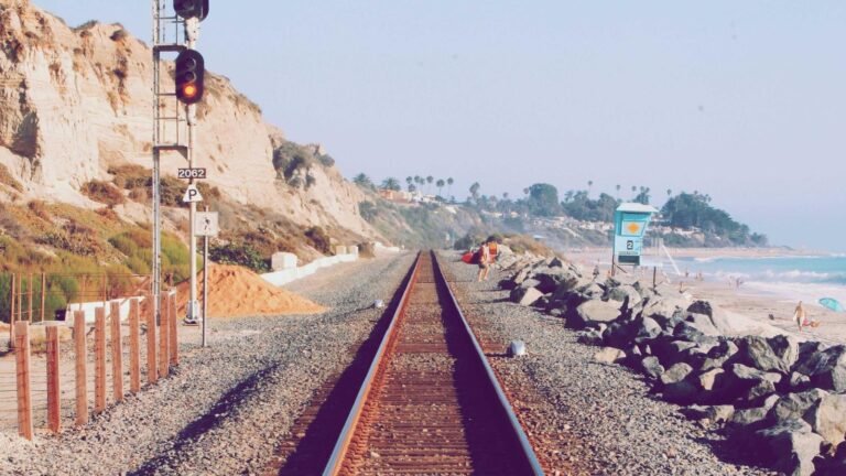 Railroad tracks along a beach with cliffs on the left, a lifeguard tower, and people surfing and sunbathing on the right.