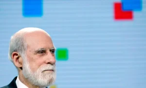 Elderly man with a white beard in profile against a blue screen with colorful square patterns.