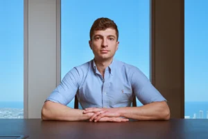 Man in a light blue shirt sits at a table with a cityscape and blue sky visible through a large window behind him.