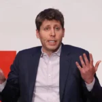 Man in a suit gestures with both hands during a speech, wearing a microphone headset, against a white and red backdrop.