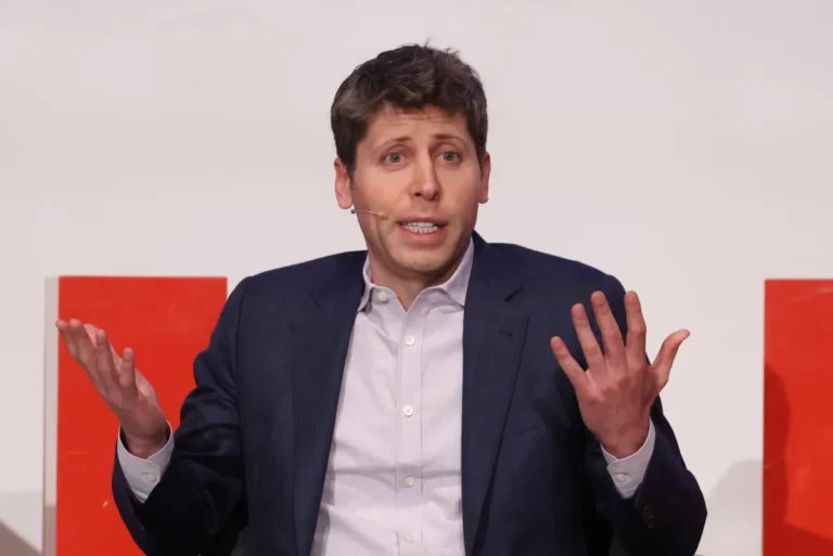 Man in a suit gestures with both hands during a speech, wearing a microphone headset, against a white and red backdrop.