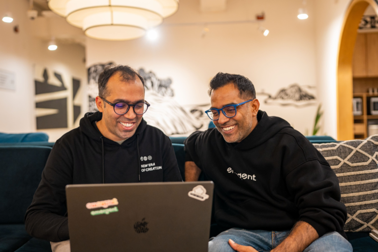 Two people smiling at a laptop in a cozy office, both wearing black hoodies, one says "new era of creation," the other "em...