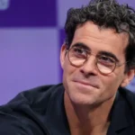 Person with glasses and curly hair in front of a purple backdrop with blurred text.