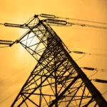 Silhouette of a tall electricity pylon against an orange sky with visible power lines.