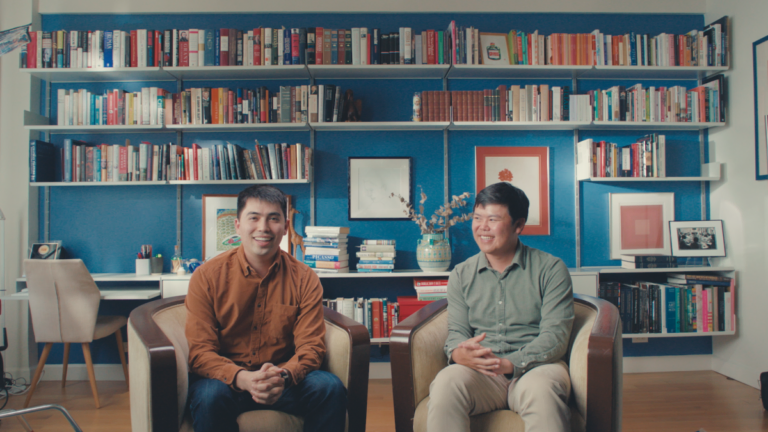Two men seated and smiling in a room with blue walls, surrounded by bookshelves filled with colorful books and framed art.