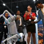 A robot interacts with a group of people taking photos at an indoor tech event, with banners and displays in the background.