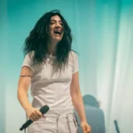 Person with long hair in a white outfit holding a microphone, smiling on stage with a teal backdrop.